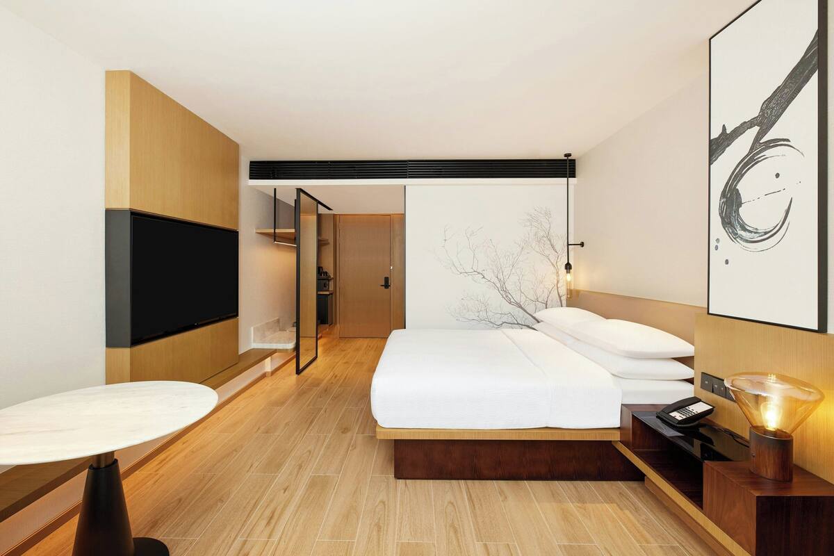 Una habitación de hotel moderna con una cama grande, un televisor de pantalla plana montado en la pared, una mesita de noche y una lámpara.
