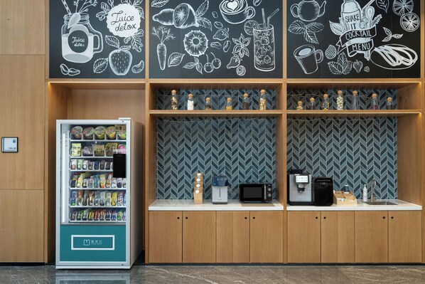 Máquina de vendas com lanches, máquina de café e micro-ondas sobre um balcão com armários de madeira e painel de backsplash em padrão herringbone.