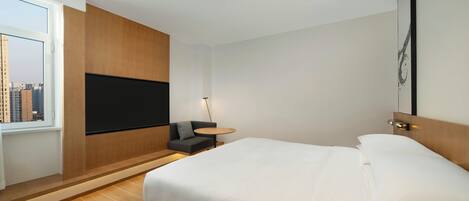 A modern hotel room with a large bed, a flat-screen TV mounted on the wall, a small round table with a lamp, and a view of the city through a large window.