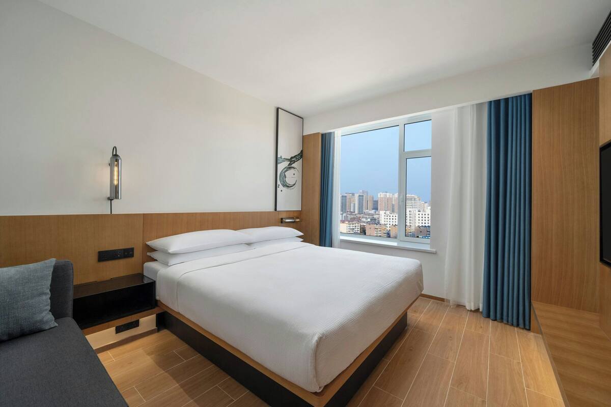 Habitación de hotel moderna con una cama grande, un sofá, una mesita de noche, una lámpara y una ventana con vista a la ciudad.