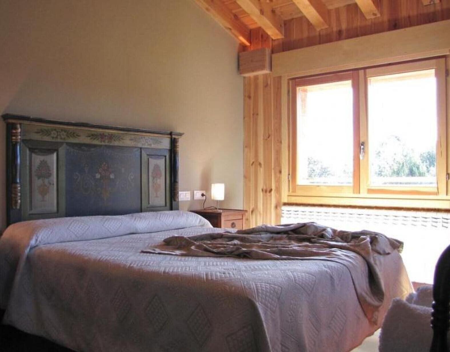 Un dormitorio con una cama, una mesita de noche, un ventanal y techo de madera.