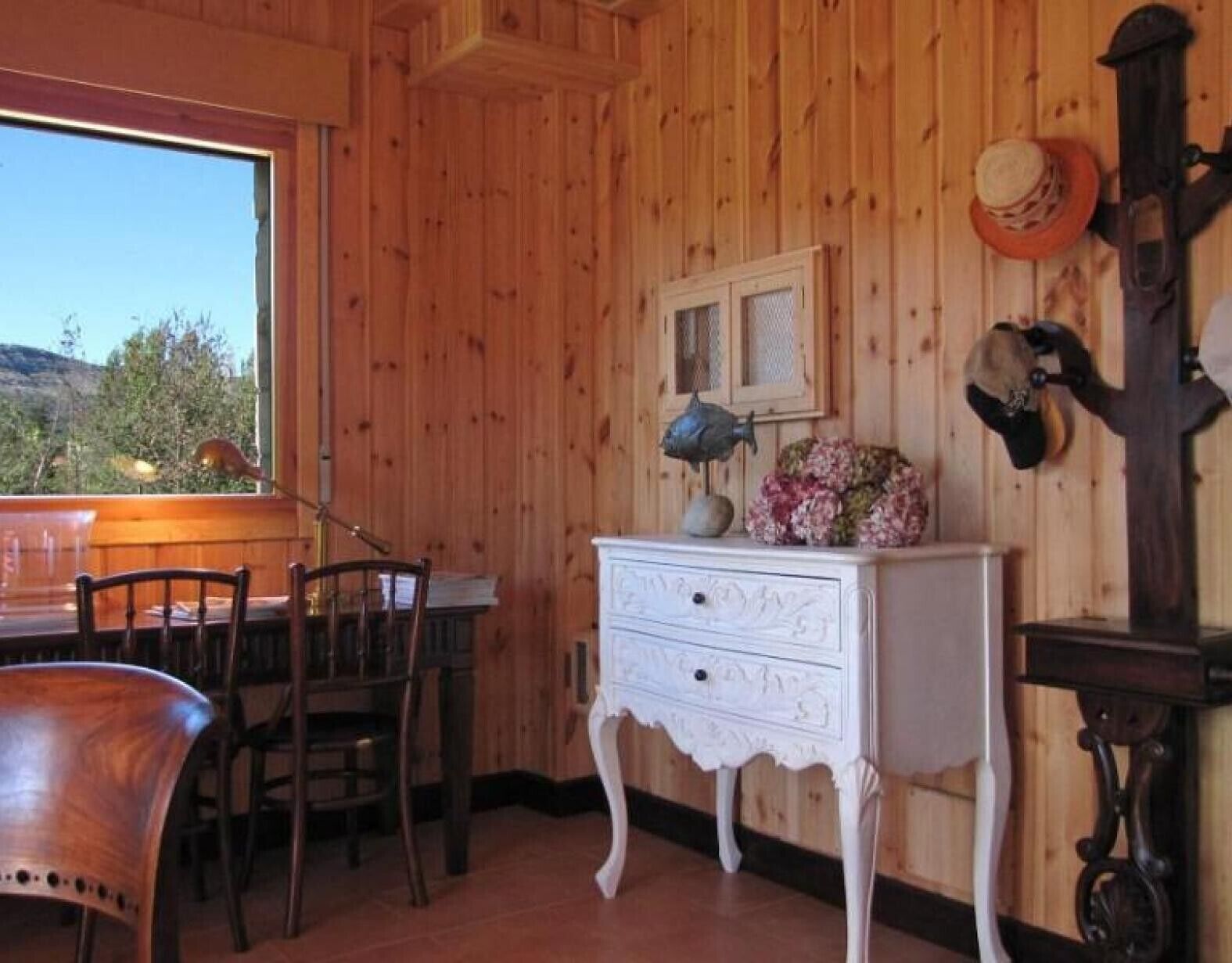 Une pièce aux murs lambrés, comprenant une table à manger, des chaises, une commode blanche et un porte-chapeaux fixé au mur.