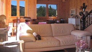 Interior de una cabaña de madera con sofá, mesa de comedor, sillas y vistas al exterior.