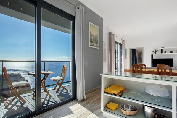 A balcony with a view of the sea, a dining area with a glass table and chairs, and a shelf with kitchenware.