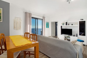 A room with a dining table, chairs, a sofa, a TV, and a view of the sea.