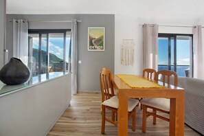 A dining area with a table and chairs, a vase, a framed picture on the wall, and a view of the sea through large windows.