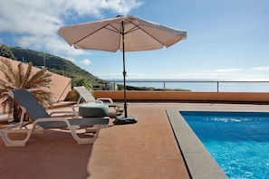 A poolside area with lounge chairs, an umbrella, and a view of the sea.