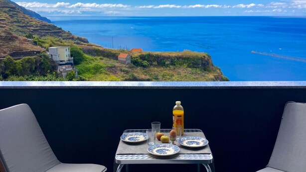 A balcony with a table set for two, overlooking a coastal landscape with a clear view of the sea and distant hills.