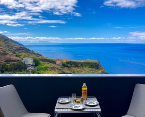 Un balcone con un tavolo apparecchiato per due, affacciato su un paesaggio costiero con vista libera sul mare e colline lontane.