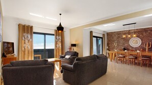 A modern living room with a sofa, a dining area with chairs, a table, and a ceiling light fixture.