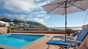 A swimming pool with lounge chairs and an umbrella, overlooking a hillside with houses.