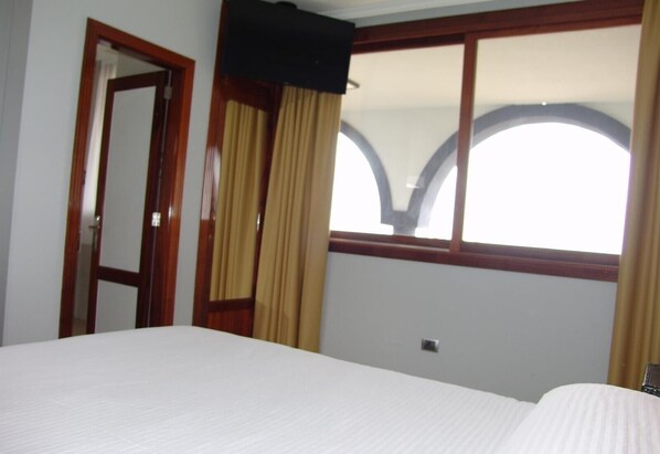 Habitación de hotel con una cama, una puerta, un ventanal con cortinas y un televisor instalado en la pared.