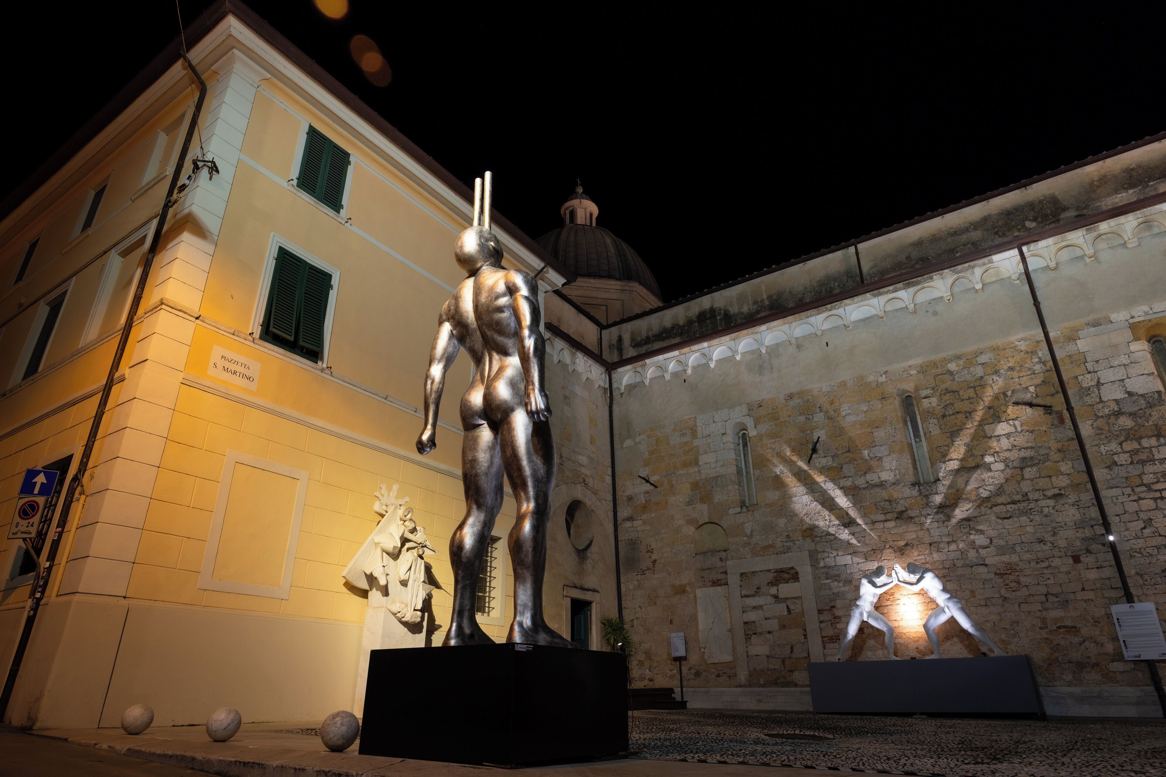 Una statua in bronzo raffigura un uomo in posa classica, una statua più piccola di una figura femminile e un rilievo che mostra due figure in combattimento, esposti all'aperto di fronte a una facciata con una cupola e finestre ad arco.