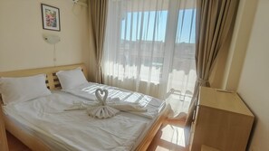 A hotel room with a bed, a bedside table, a wall-mounted lamp, and a window with curtains. - Apartment - Sleeps 6, Pet Friendly & 6 Pools (Sunny beach)