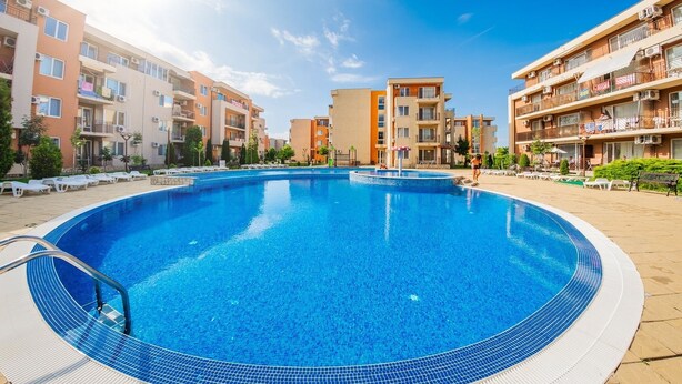 A swimming pool surrounded by multi-story residential buildings with balconies, a clear sky, and a few lounge chairs.
