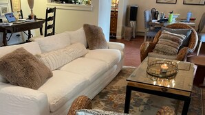 A living room with a white sofa, wicker chairs, a coffee table with a candle, and a kitchen in the background.