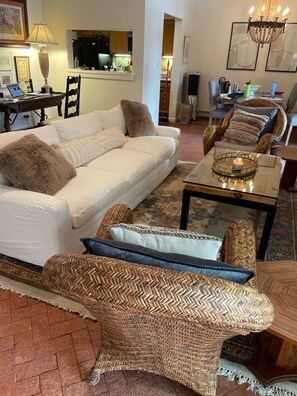 Ein Wohnzimmer mit einem weißen Sofa, Korbstühlen, einem Couchtisch mit Kerze und einer Küche im Hintergrund.