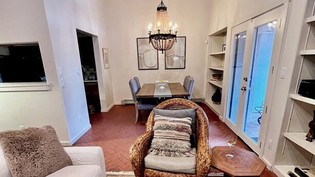 A cozy living room with a sofa, wicker chair, dining table, chandelier, and a view through a door.