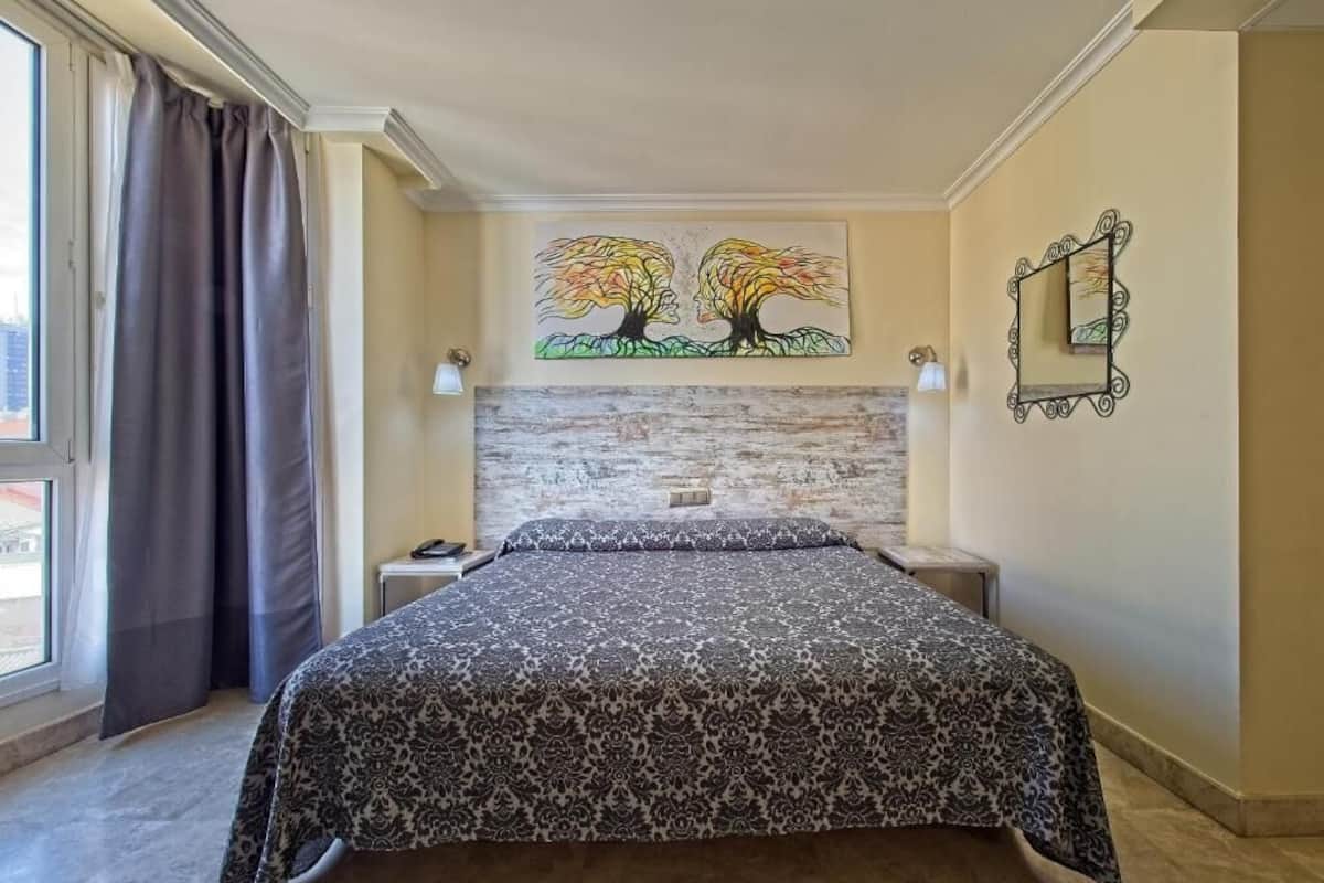 A bedroom with a bed, a headboard, a painting of trees, a mirror, and a window with curtains.
