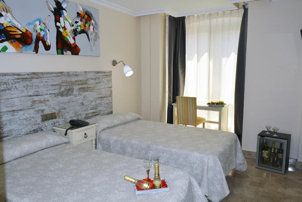 Una camera d'albergo con due letti, un tavolino con una bottiglia e bicchieri, un quadro appeso al muro e vista all'esterno da una finestra.