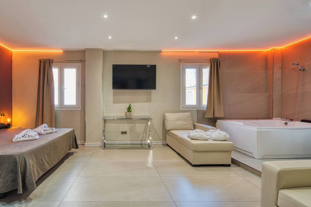 A modern hotel room with a bed, a sofa, a small table with a plant, a TV, and a bathtub.