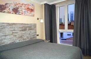 Una camera d'albergo con un letto, un quadro, una lampada e vista sui palazzi attraverso una finestra.