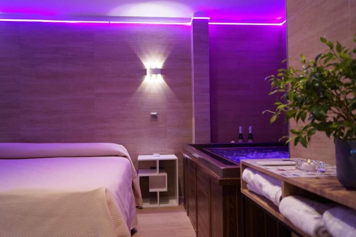A modern spa room with a bed, a massage table, a wooden cabinet with bottles, and a small table with towels and a plant.
