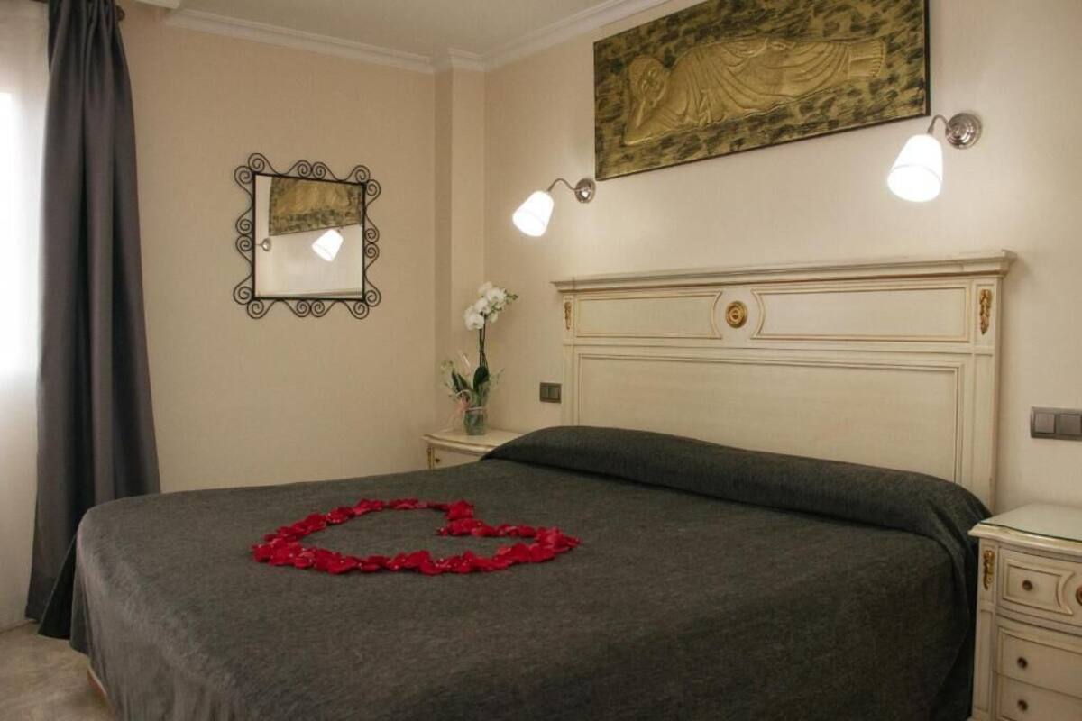 A hotel room with a bed, a nightstand, a mirror, and a wall-mounted light fixture.
