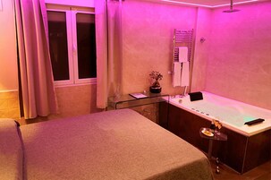 Ein Spa-Raum mit einer Massageliege, einem kleinen Tisch mit Flasche und Glas, einer Topfpflanze und einem Fenster mit Vorhängen.
