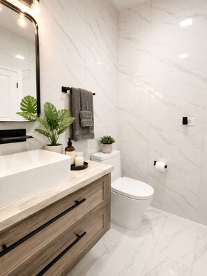 A modern bathroom with a marble floor, a wooden vanity, a white toilet, and a mirror with lights.