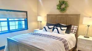 Un dormitorio con una cama de madera, dos mesitas de noche con lámparas, una ventana con persianas y un arreglo de guirnaldas colgado en la pared.