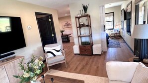Un salón espacioso con televisor de pantalla plana, una mesa de centro de madera y un sofá beige. Se aprecia una estantería con adornos y una silla al fondo.