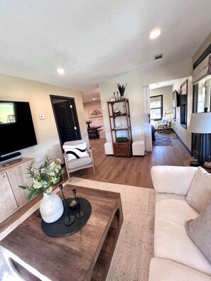 Een ruime woonkamer met een flatscreen televisie, een houten salontafel en een beige bank. Achterin staat een boekenplank met decoratieve objecten en een stoel.