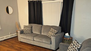 A living room with a grey sofa, a round mirror, and a window with black curtains.