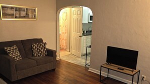 A living room with a sofa, a TV on a stand, and a kitchen visible through an open door.
