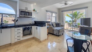 A modern kitchen with white cabinets, a black countertop, and a built-in oven. There is a glass dining table with chairs, a sofa, and a TV in a balcony area with a view of palm trees and the ocean. - Beachfront Escape at Mykonos Bay Resort 401B (San José del Cabo)