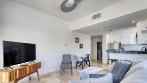 A modern living room with a flat-screen TV, wooden console table, grey sofa, and a dining area with chairs. - Beachfront Escape at Mykonos Bay Resort 401B (San José del Cabo)