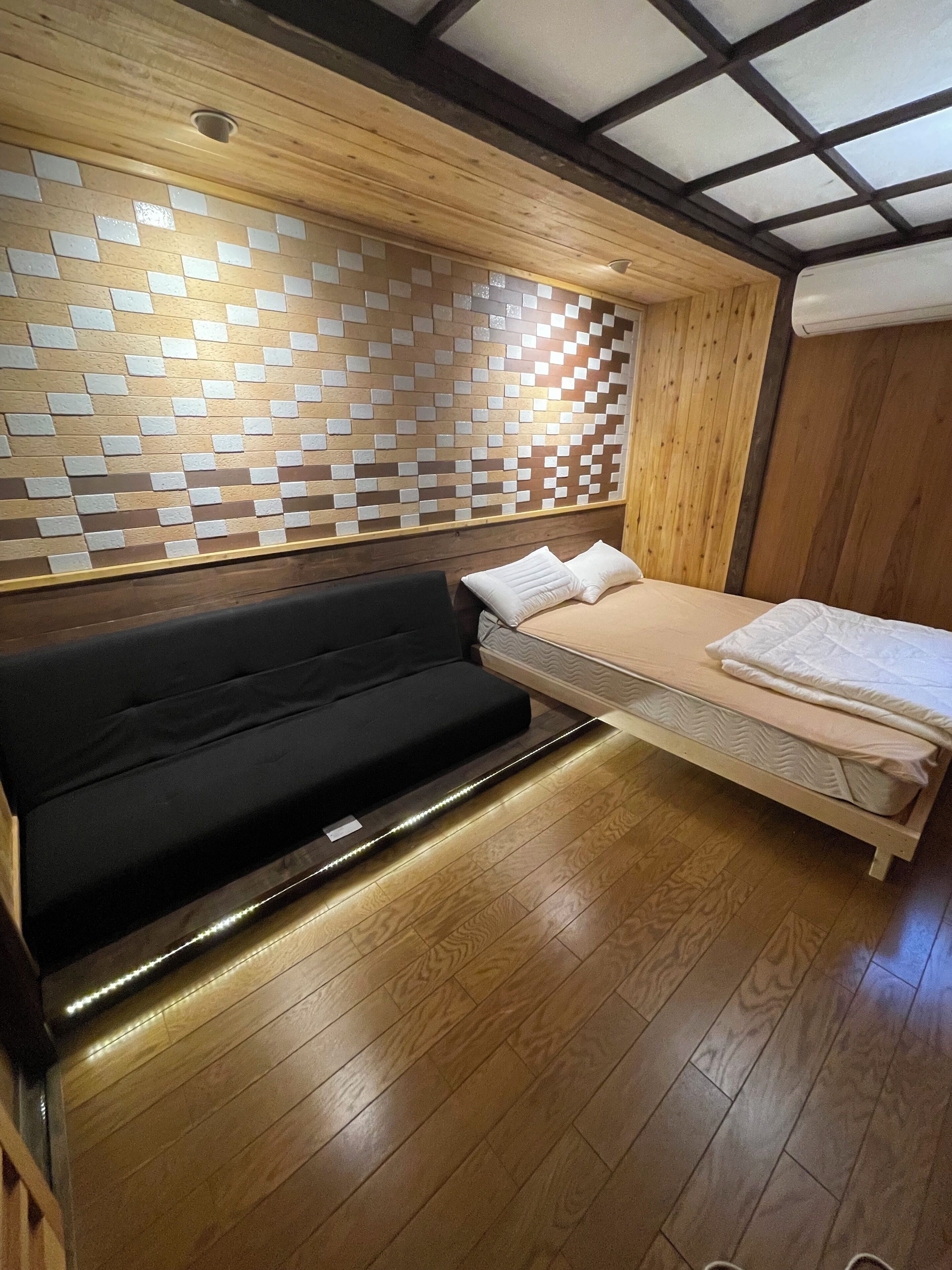 Une chambre moderne avec un plafond en bois, un mur orné de motifs géométriques en carreaux, un canapé noir et un lit avec une literie blanche.