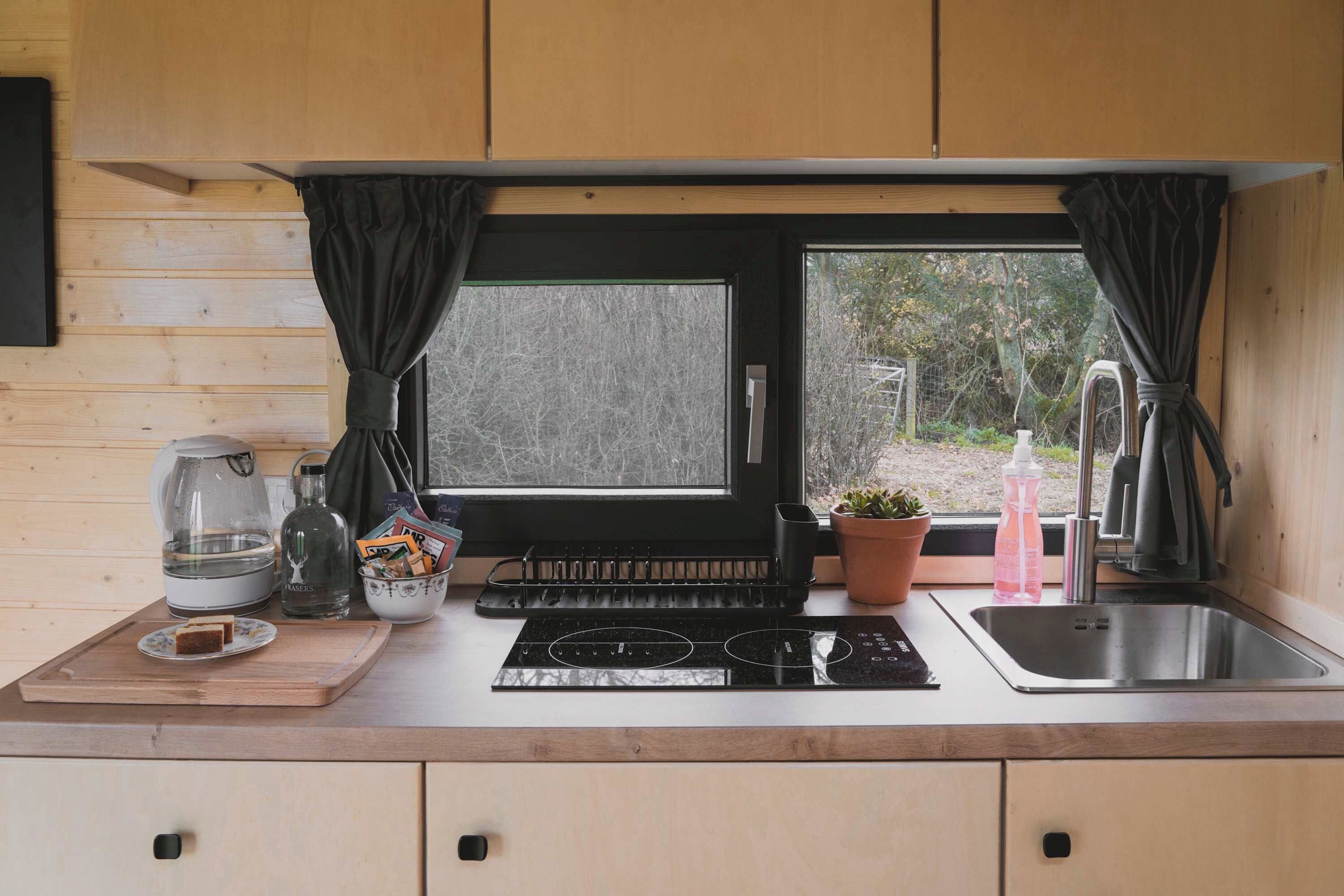 Una cocina compacta con ventana, hervidor, botella, tabla de cortar y lavamanos.