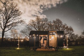 A small wooden cabin with a porch, a bench, and string lights, surrounded by trees under a starry sky. - The Field View 2 by Tiny Away (Egerton)