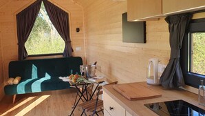 A cozy cabin interior with a green sofa, a small dining table, a kitchen area, and a window with curtains.