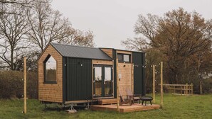 A small wooden cabin with a sloped roof, large windows, and a deck with outdoor seating.