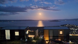 Una vista de un lago al anochecer con la luna reflejada en el agua, rodeado de pequeñas casas con ventanas visibles y algunas luces exteriores.