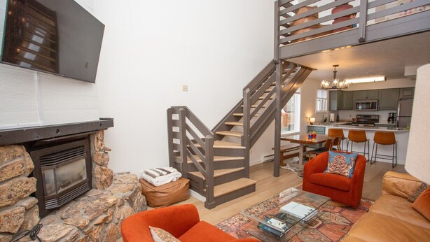 Amplia sala de estar con chimenea de piedra, una escalera de madera y una cocina al fondo.