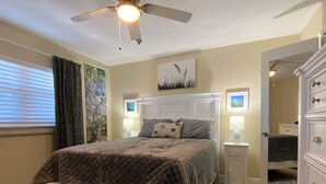 Une chambre à coucher avec un lit queen-size, deux tables de chevet avec des lampes, un ventilateur de plafond, une grande fenêtre avec des stores et une porte avec un miroir.
