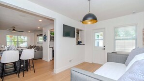 Un salon moderne avec un canapé gris, un coin repas avec une table et des chaises, une cuisine en arrière-plan et un ventilateur de plafond.