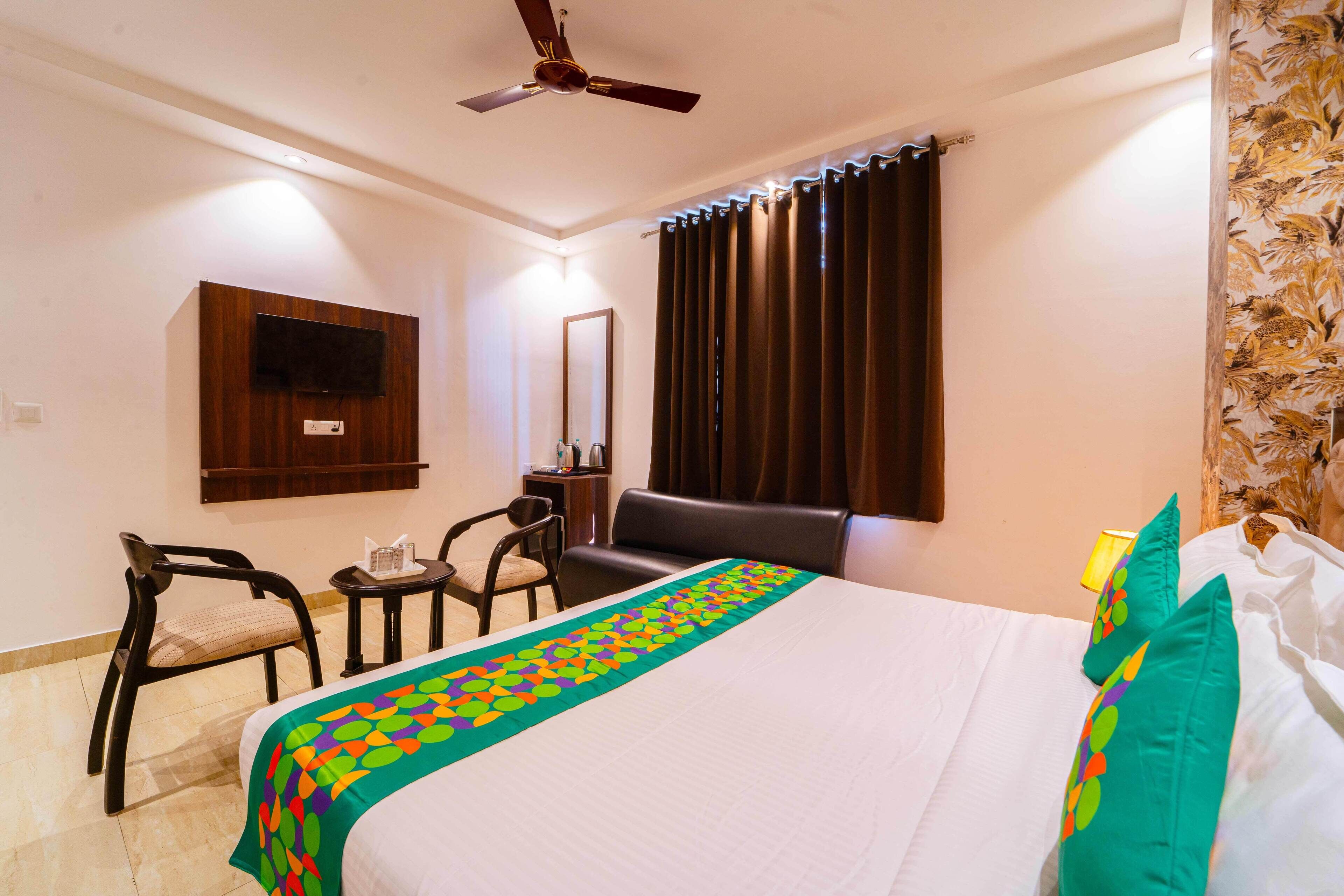 A hotel room with a bed, a TV mounted on the wall, a ceiling fan, a desk with a chair, and a window with curtains.