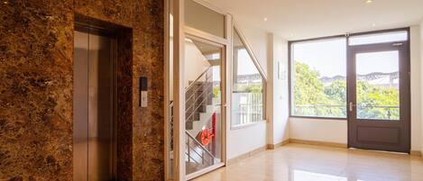 A modern interior with a marble wall, an elevator, a staircase with glass railings, and a large window offering a view of greenery and a bridge.