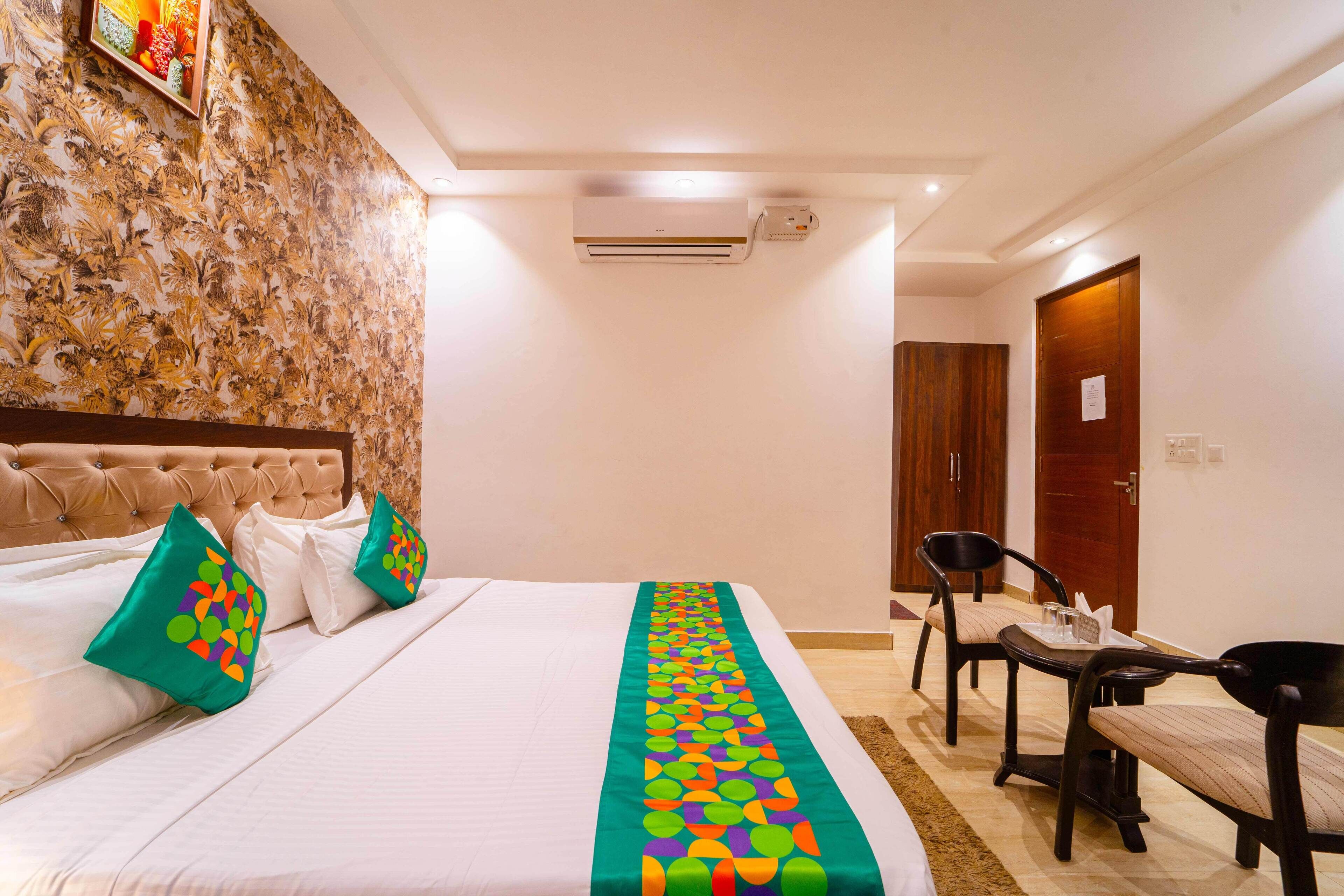 A hotel room with a large bed, a headboard, a wall with a floral pattern, an air conditioning unit, a wooden door, and a small table with chairs.