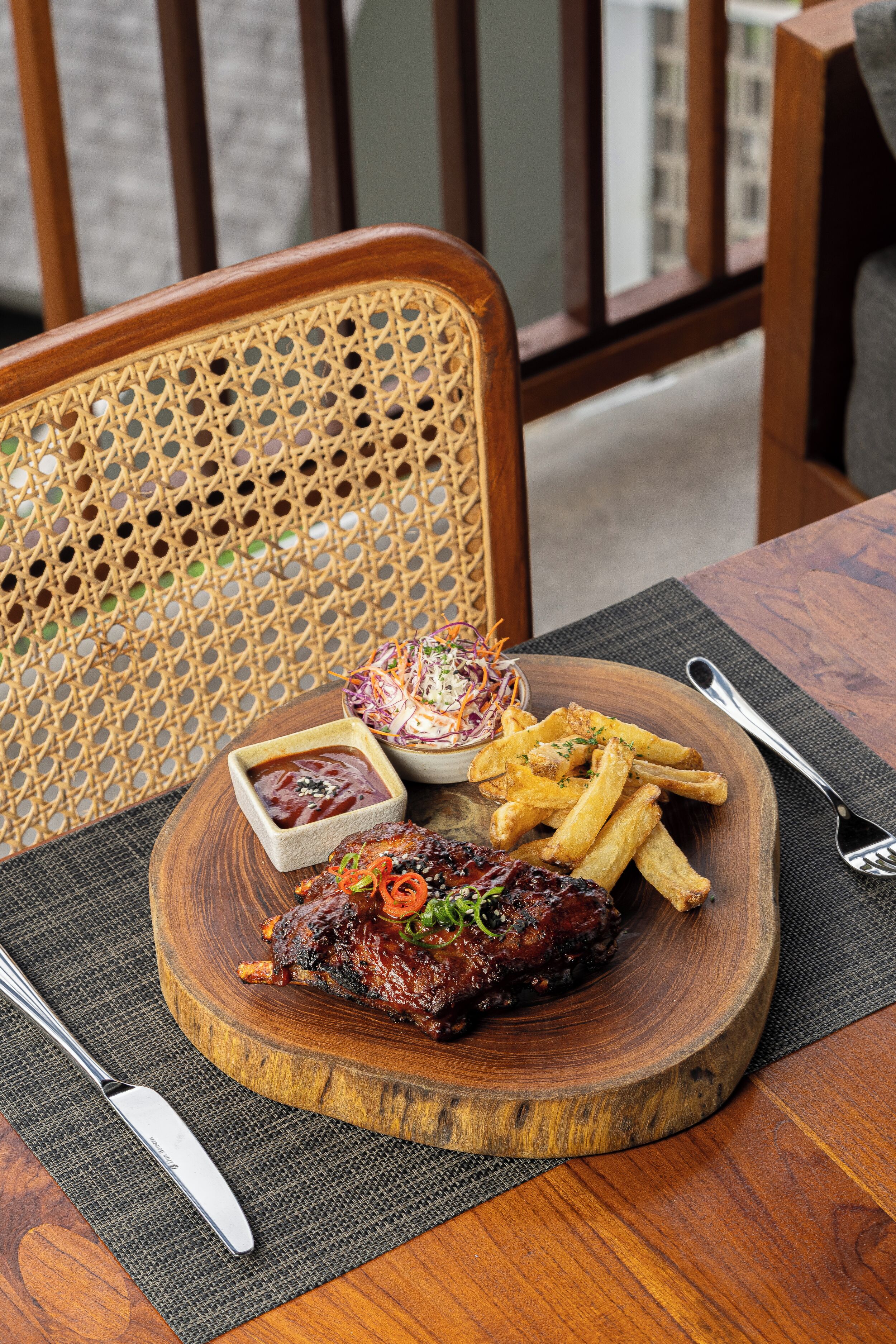 A wooden serving board with a grilled meat dish, French fries, and a side salad on a table with a woven chair and a metal fork.